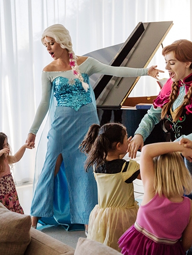 Două animatoare în costume Elsa și Anna interacționează cu copii într-un interior.